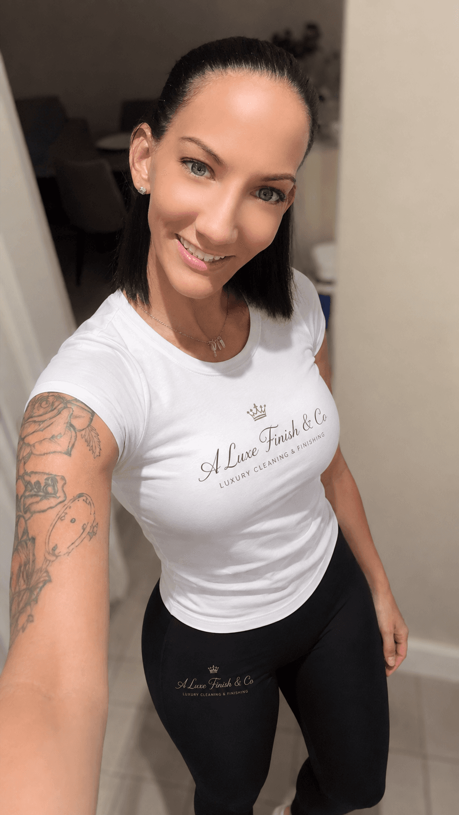 Smiling woman with dark hair and arm tattoo wearing branded white t-shirt and black leggings.
